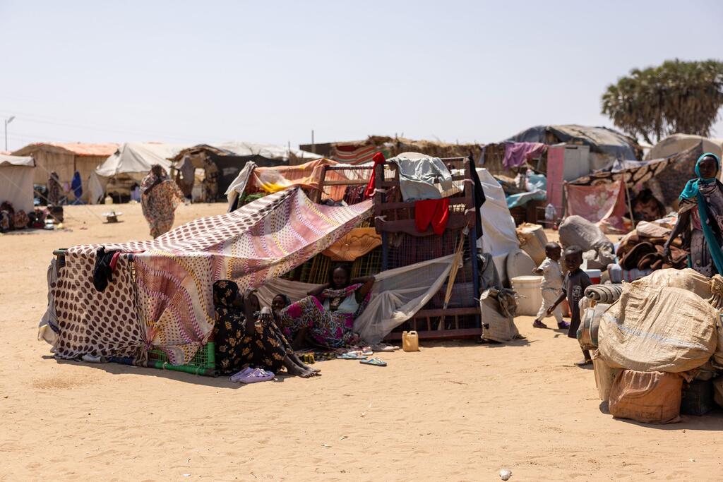 Des familles déplacées vivent dans des abris temporaires sur le site de Goz Alsalam au Soudan. Des femmes et des enfants sont assis à l'extérieur d'une tente improvisée en tissu à motifs, tandis que d'autres se promènent dans le campement de sable.