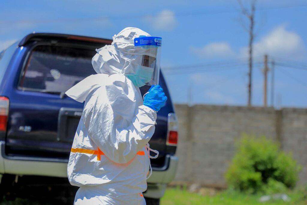 Un travailleur de santé vêtu d'une combinaison de protection blanche, d'un bouclier facial et de gants bleus se tient à l'extérieur près d'un véhicule bleu.