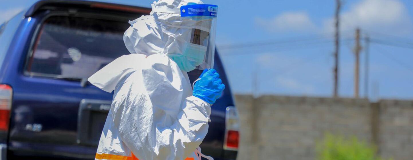 Un travailleur de santé vêtu d'une combinaison de protection blanche, d'un bouclier facial et de gants bleus se tient à l'extérieur près d'un véhicule bleu.