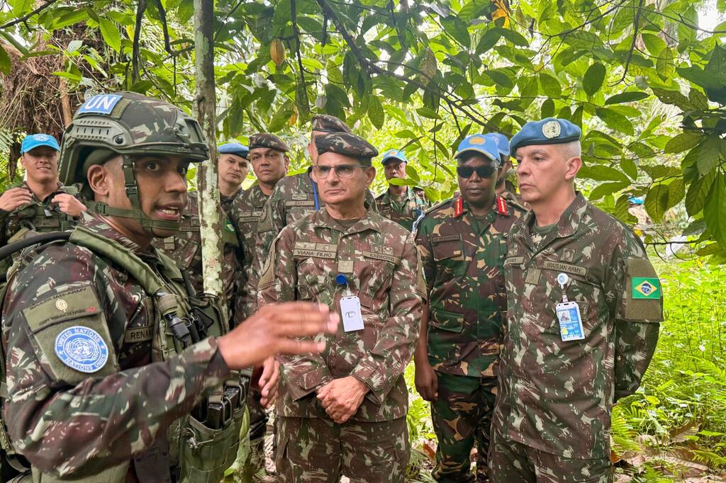 A group of UN peacekeepers in camouflage uniforms and blue berets listen to a briefing in a forest.