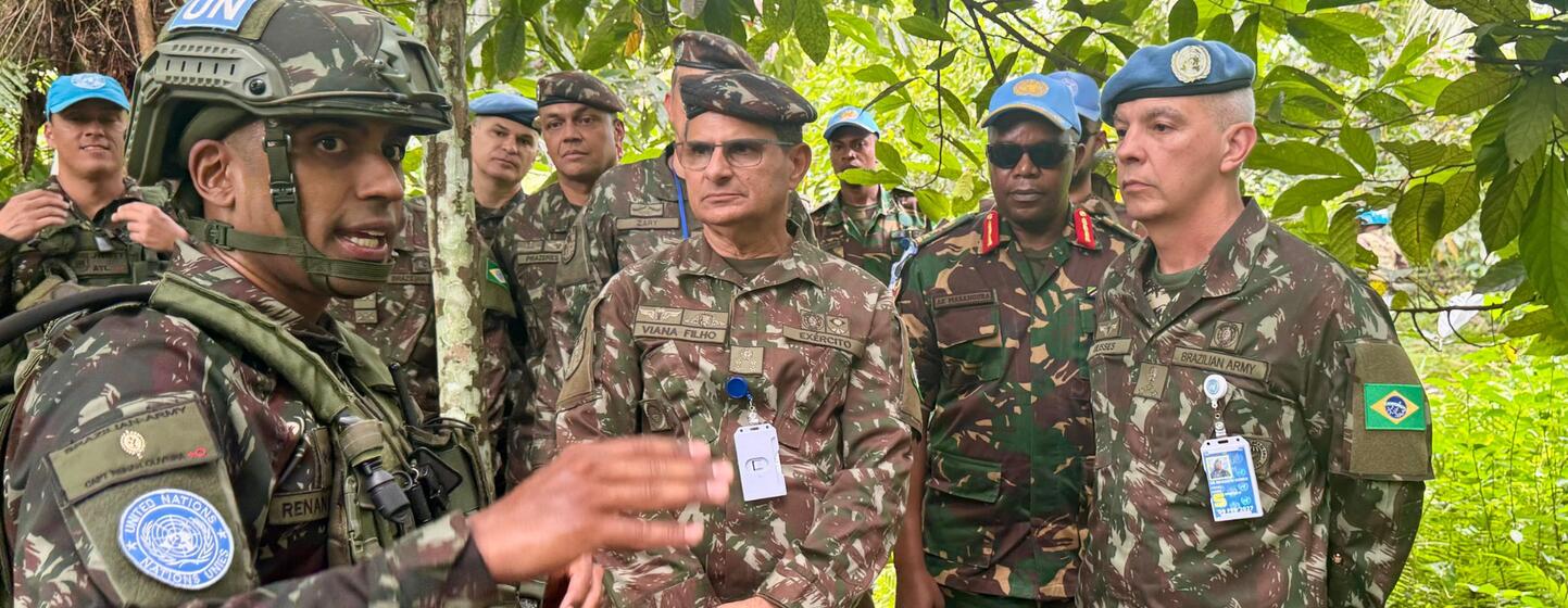Um grupo de soldados da ONU com uniformes camuflados e boinas azuis ouve um briefing em uma floresta.