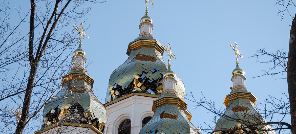 Поврежденные луковичные купола белой православной церкви в Харькове, Украина, с разбитыми медными черепицами и открытыми интерьерами, обрамленные голыми ветвями деревьев на фоне голубого неба.