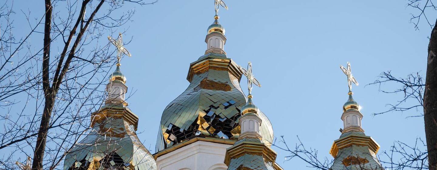 Damaged onion domes of a white Orthodox church in Kharkov, Ukraine, with broken copper tiles and exposed interiors, framed by bare tree branches against a blue sky.