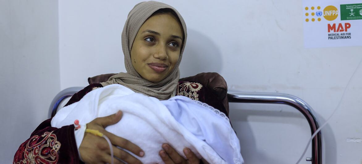 Una mujer, identificada como Rana, sosteniendo a su bebé recién nacido envuelto en una manta blanca dentro de una habitación del hospital. Ella lleva un hijab beige y una túnica de patrón, con una línea médica IV visible en su muñeca. Un logotipo de UNFPA y MAP (Ayuda Médica para los Palestinos) es visible en la pared detrás de ella.
