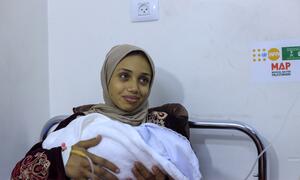 Una mujer, identificada como Rana, sosteniendo a su bebé recién nacido envuelto en una manta blanca dentro de una habitación del hospital. Ella lleva un hijab beige y una túnica de patrón, con una línea médica IV visible en su muñeca. Un logotipo de UNFPA y MAP (Ayuda Médica para los Palestinos) es visible en la pared detrás de ella.