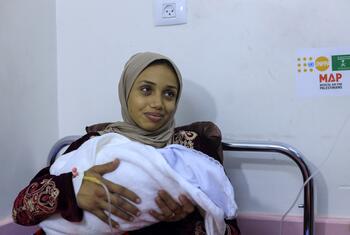 Una mujer, identificada como Rana, sosteniendo a su bebé recién nacido envuelto en una manta blanca dentro de una habitación del hospital. Ella lleva un hijab beige y una túnica de patrón, con una línea médica IV visible en su muñeca. Un logotipo de UNFPA y MAP (Ayuda Médica para los Palestinos) es visible en la pared detrás de ella.