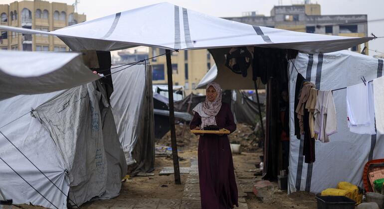 Una mujer vestida de hijab y un vestido púrpura largo camina frente a una tienda de campaña en un campamento de refugiados, llevando una bandeja.