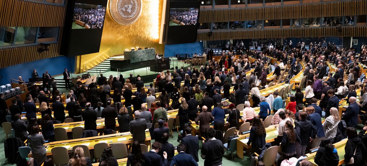 A large crowd stands in solemn silence at the United Nations General Assembly hall during a Holocaust commemoration event, honoring victims with prayers and a moment of reflection.