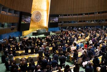 A large crowd stands in solemn silence at the United Nations General Assembly hall during a Holocaust commemoration event, honoring victims with prayers and a moment of reflection.