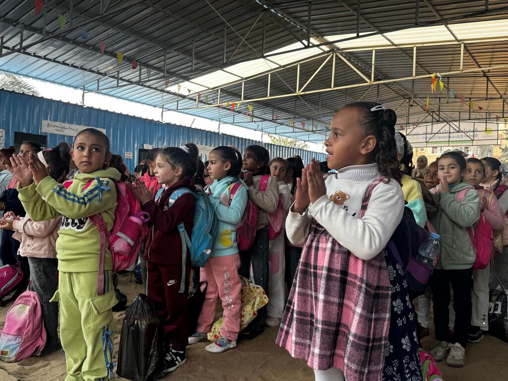 Kikundi cha watoto wa Palestina wamesimama katika kituo cha muda cha kujifunza huko Gaza, wakipiga makofi. Wanavaa mavazi yenye rangi na mkoba, na ishara za UNICEF na maandishi ya Kiarabu zinaonekana kwenye mandhari ya nyuma.