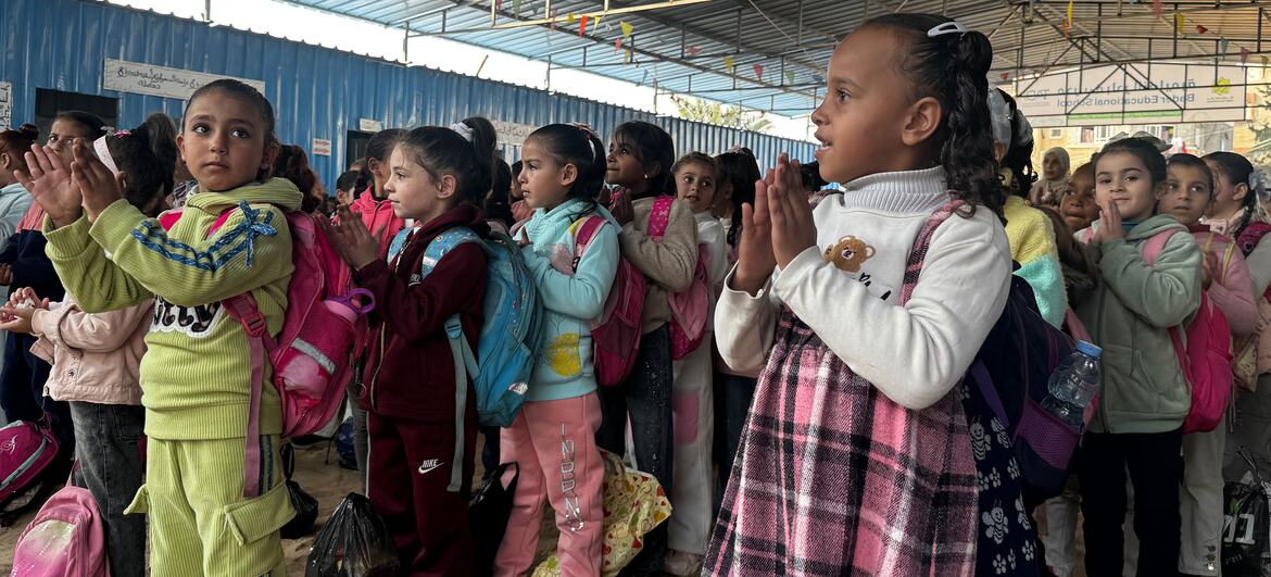 Um grupo de crianças palestinas está em pé em um centro de aprendizagem temporário em Gaza, aplaudindo, vestindo roupas coloridas e mochilas, com sinais do UNICEF e texto árabe visíveis no fundo.