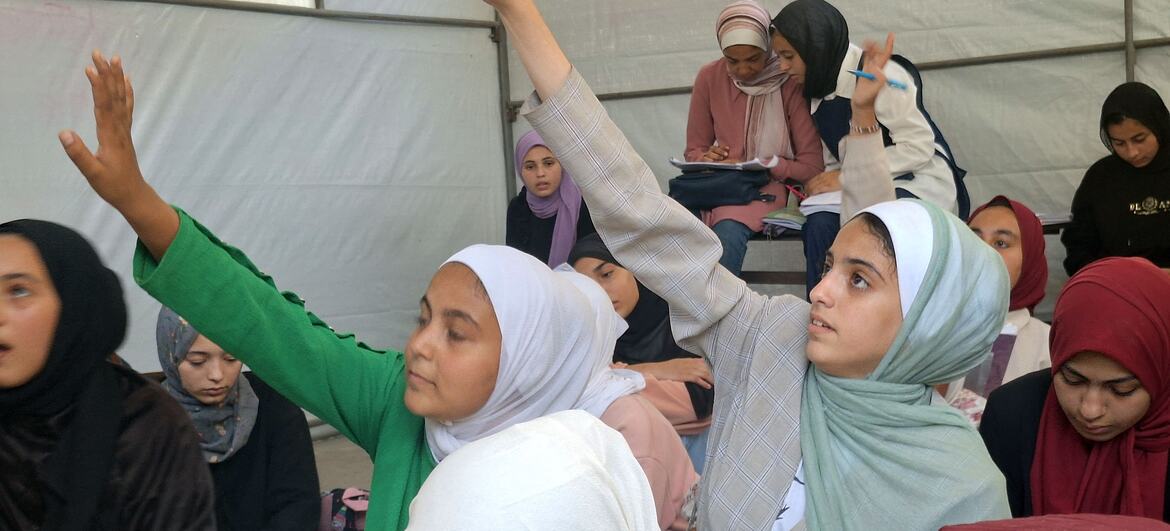 Los niños levantan las manos para responder preguntas en un centro de aprendizaje apoyado por UNICEF en la Escuela Aeshyia en Deir al-Balah, Gaza, estudiando bajo tiendas de campaña en medio de infraestructuras dañadas.