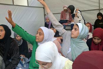 Los niños levantan las manos para responder preguntas en un centro de aprendizaje apoyado por UNICEF en la Escuela Aeshyia en Deir al-Balah, Gaza, estudiando bajo tiendas de campaña en medio de infraestructuras dañadas.