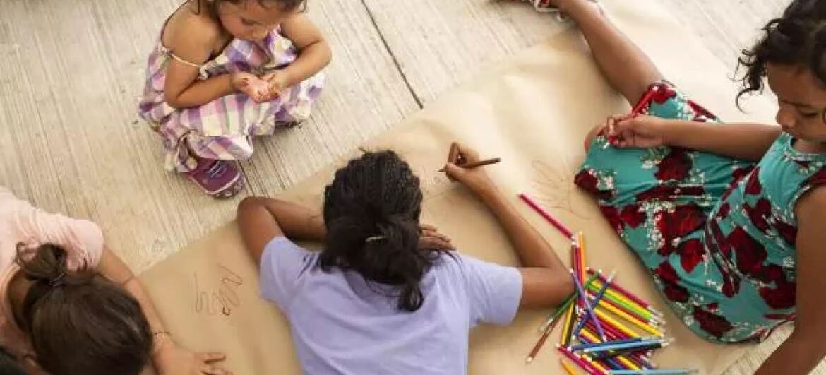 Um grupo de jovens adolescentes envolvidos em uma atividade de apoio psicosocial baseada em artes, desenhando em uma grande folha de papel com lápis coloridos enquanto sentados no chão.