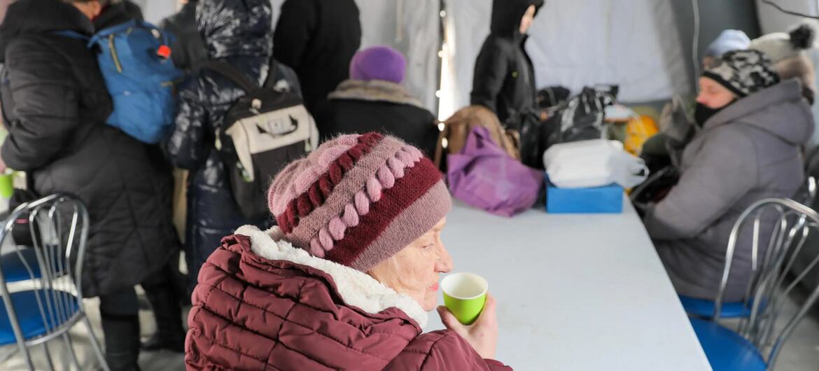 Пожилая женщина, одетая в зимнюю шляпу и пальто, выпивает из зеленой чашки на столе в лагере беженцев, на фоне которого стоят другие беженцы.