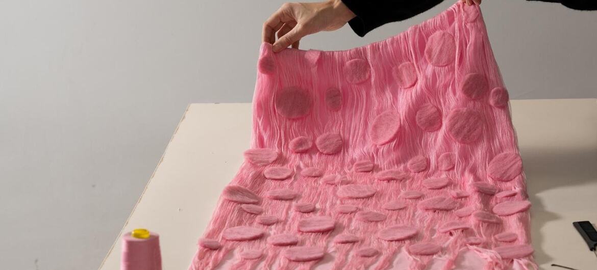 Uma pessoa segura um pedaço de tecido rosa com padrões de textura circular, com uma bobina de fio rosa e uma fita de medição amarela em uma mesa branca.