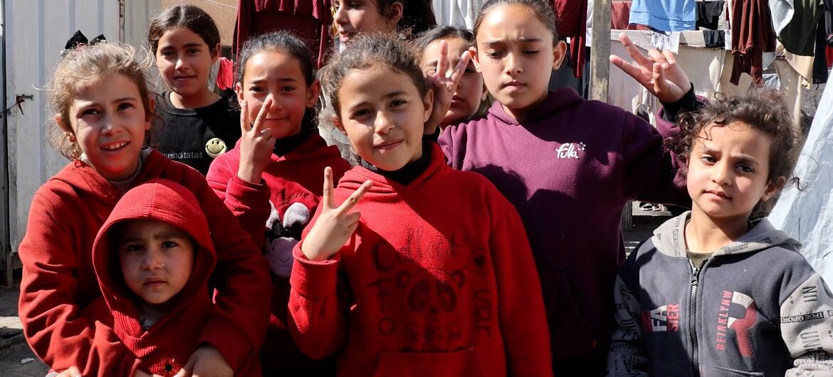 مجموعة من الأطفال الفلسطينيين يقفون بين خيام النازحين في مدينة غزة، بعضهم يبتسم ويرفع يده بعلامة السلام، تظهر في الخلفية ملابس معلقة بين الخيام.