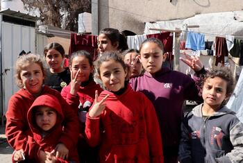 مجموعة من الأطفال الفلسطينيين يقفون بين خيام النازحين في مدينة غزة، بعضهم يبتسم ويرفع يده بعلامة السلام، تظهر في الخلفية ملابس معلقة بين الخيام.