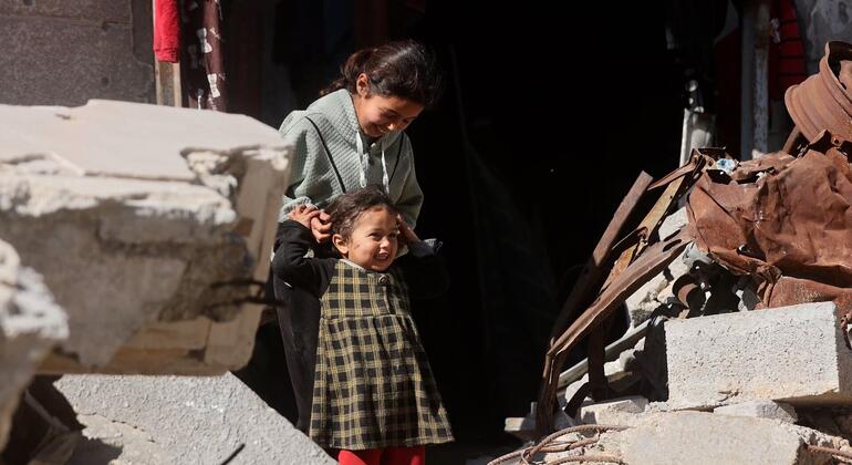 طفلتان فلسطينيتان تقفان بين أنقاض خرسانية وهياكل معدنية ملتوية في مدينة غزة. تبدوان مبتسمتين وهما تلعبان تحت أشعة الشمس.