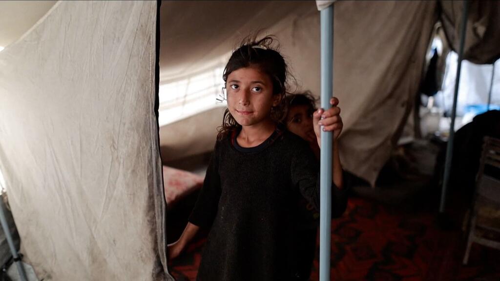 Une jeune fille palestinienne se tient à l'intérieur d'un refuge de tente improvisé, tenant un poteau de soutien et regardant vers la caméra.