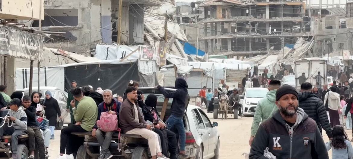 Un grupo de personas desplazadas en Gaza, incluidos hombres, mujeres y niños, viajan en un carro improvisado tirado por un automóvil a través de una calle bordeada de edificios destruidos y refugios improvisados.