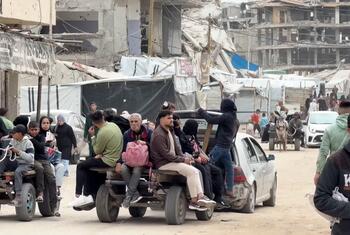 गाजा में विस्थापित लोगों का एक समूह, जिसमें पुरुष, महिलाएं और बच्चे शामिल हैं, एक कार द्वारा खींची गई अस्थायी गाड़ी में एक सड़क के माध्यम से यात्रा करते हैं, जो ध्वस्त इमारतों और अस्थायी आश्रयों से घिरी हुई है।