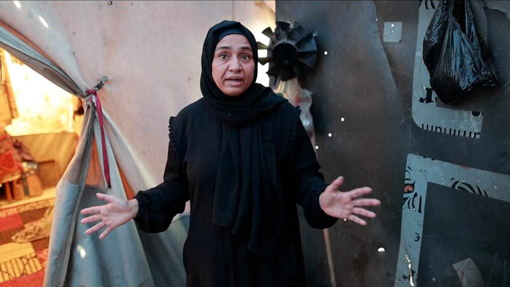 صورة لفتاة فلسطينية ترتدي حجابًا أسودًا وعباء، واقفة بأيدي مُمتدة أمام ملجأ خيمة مؤقت في غزة.
