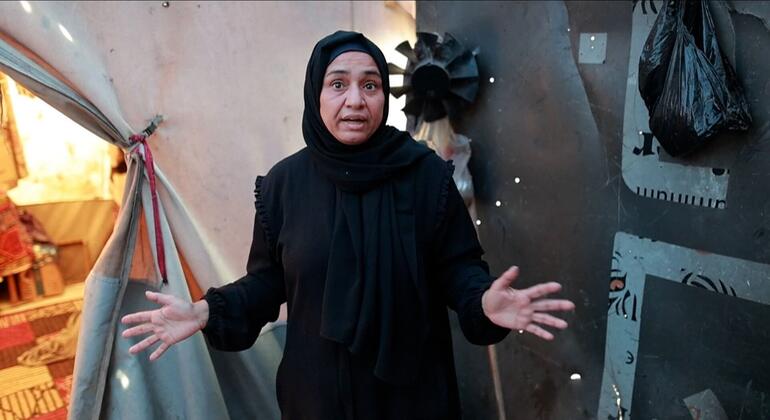 صورة لفتاة فلسطينية ترتدي حجابًا أسودًا وعباء، واقفة بأيدي مُمتدة أمام ملجأ خيمة مؤقت في غزة.