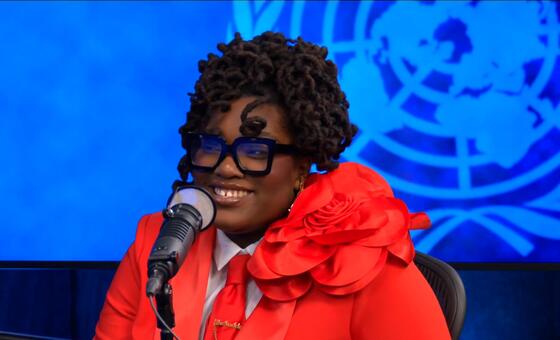 Une femme afro-américaine souriante aux cheveux bouclés et aux lunettes, vêtue d'un costume rouge avec une grande rose rouge, parle dans un microphone sur fond bleu avec le logo des Nations Unies.