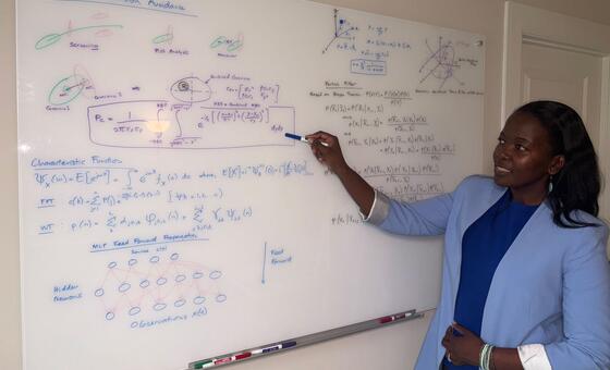 Alinda Mashiku, an aerospace engineer and NASA Program Manager, stands beside a whiteboard filled with complex mathematical equations and diagrams related to satellite collision avoidance and orbital mechanics, as she explains the concepts.