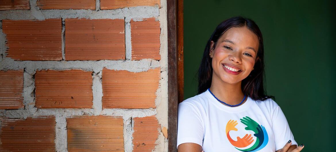 Uma jovem brasileira sorridente, de cabelos escuros, está de pé com os braços cruzados ao lado de uma parede de tijolos. Ela veste uma camiseta branca com o logotipo da UNICEF, relacionada a uma campanha de vacinação contra o HPV no Brasil.