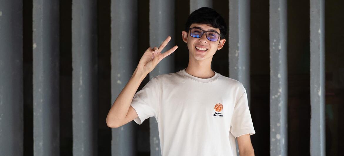 Um adolescente brasileiro sorridente, usando óculos e uma camiseta branca, faz um sinal de paz, promovendo a campanha de vacinação contra o HPV da UNICEF no Brasil.