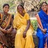 Três mulheres indianas sorridentes, vestindo saris tradicionais coloridos, sentam-se juntas ao ar livre sobre um muro de pedra.