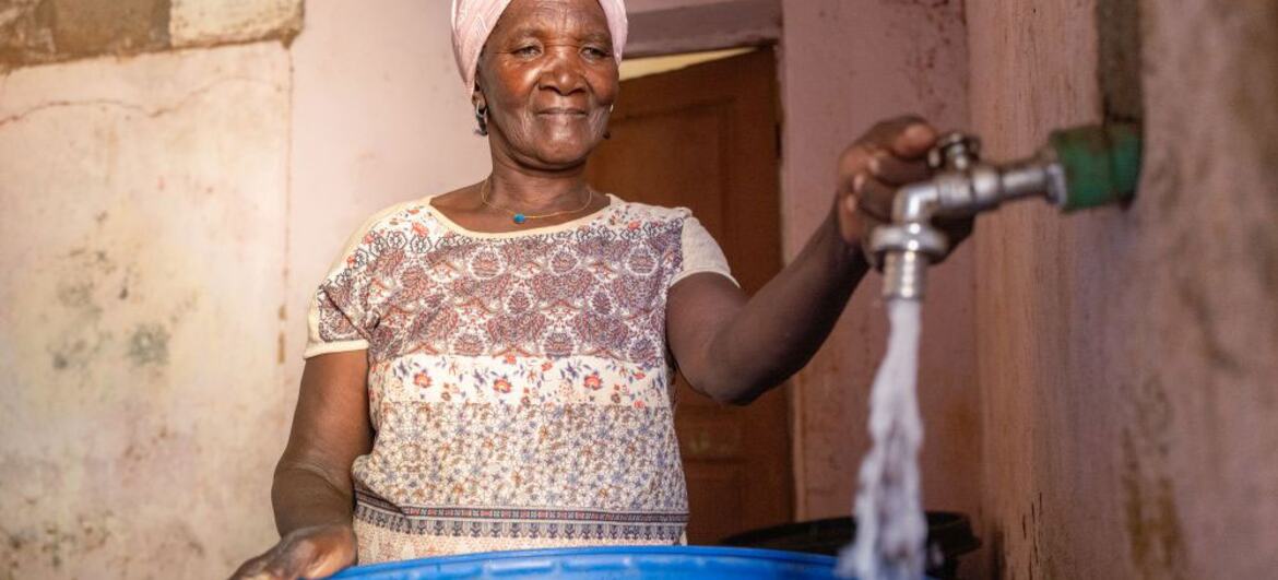 Uma mulher em Cabo Verde enche um barril azul com água de uma torneira, simbolizando melhoria da irrigação e desenvolvimento rural que permite que as famílias permaneçam e construam futuros em suas comunidades.