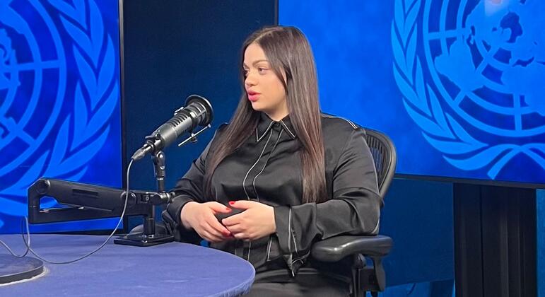 Наталия Томенко, выжившая из Холокоста, сидит на собеседовании UN News, говоря в микрофоне перед эмблемой ООН.