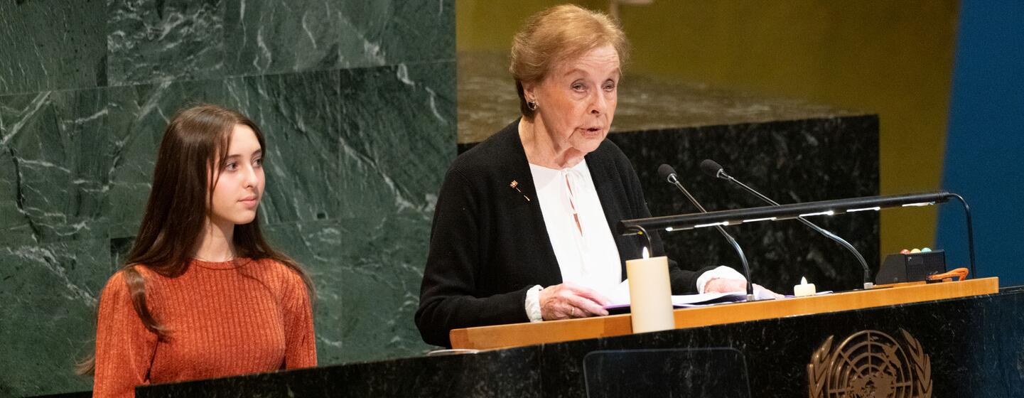 An elderly Holocaust survivor speaks at a United Nations podium during a Holocaust remembrance ceremony, with a young woman standing beside her.