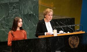 An elderly Holocaust survivor speaks at a United Nations podium during a Holocaust remembrance ceremony, with a young woman standing beside her.