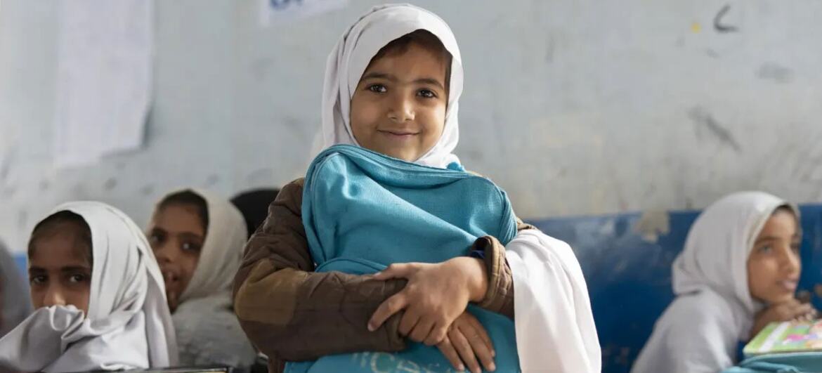 بلوچستان، پاکستان میں ایک کلاس روم میں ایک نوجوان لڑکی جو یونیسف کا بیگ تھام کر مسکرا رہی ہے۔ وہ دوسرے طالب علموں کے ساتھ گھیر لی گئی ہے، جس کی دیوار پر یونیسف اور جی پی ای کے لوگو نظر آتے ہیں۔
