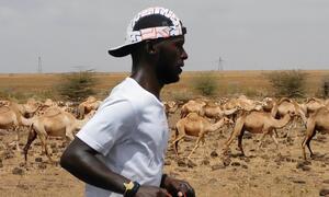 Mwanariadha wa kiume aliyevaa shati na kofia nyeupe anakimbia kupitia eneo kavu na kavu lenye ngamia kwenye mandhari ya nyuma, wakiwakilisha mandhari za uhamiaji.