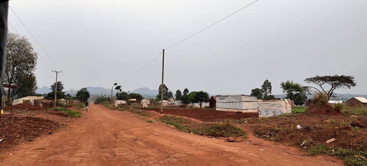 A panoramic view of a rural landscape in Byali, Uganda, featuring a wide dirt road with makeshift structures on either side, sparse trees, and mountains in the distance under an overcast sky.
