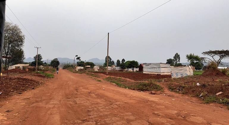 منظر بانورامي لمنظر ريفي في بيالي بأوغندا، يضم طريقًا ترابيًا واسعًا مع هياكل مؤقتة على جانبيه، وأشجارًا متفرقة، وجبالًا في الأفق تحت سماء غائمة.