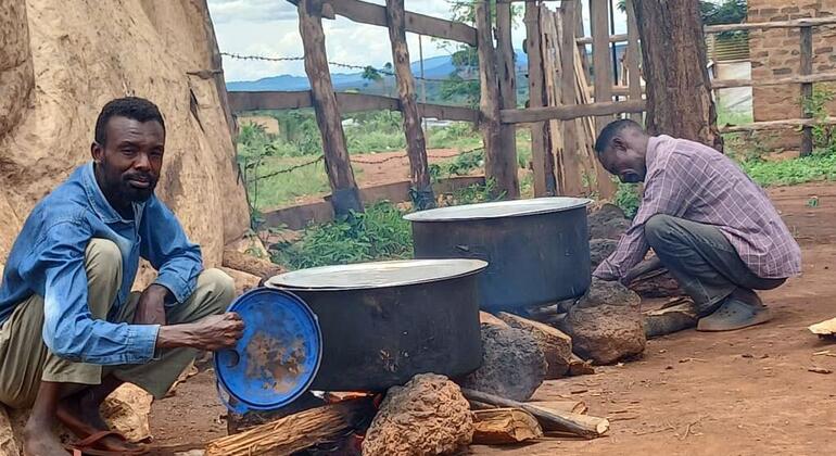 اثنان من الرجال الأفريقيين يطبخون في الهواء الطلق في أوغندا أو دارفور. رجل في قميص أزرق يلتصق بجوار وعاء كبير فوق نار مفتوحة، بينما رجل آخر في قميص مزدوج يهتم بعاء ثاني.