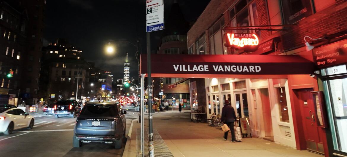 Night view of the Village Vanguard jazz club in Greenwich Village, New York City, with its red neon sign and awning, and the Manhattan skyline visible in the distance.