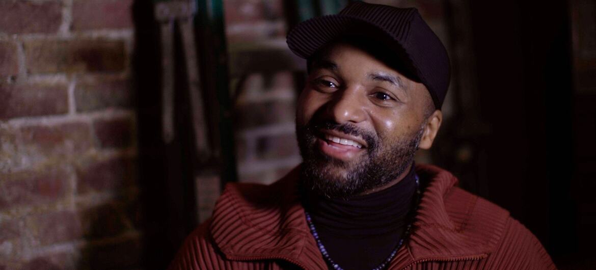 A smiling African American man with a beard wearing a black cap, black turtleneck, and a red jacket, standing in front of a brick wall.