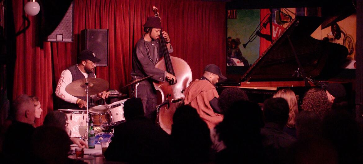 Um trio de jazz se apresentando no palco do Village Vanguard, com um baixista, um baterista e um pianista, com o público assistindo em primeiro plano.