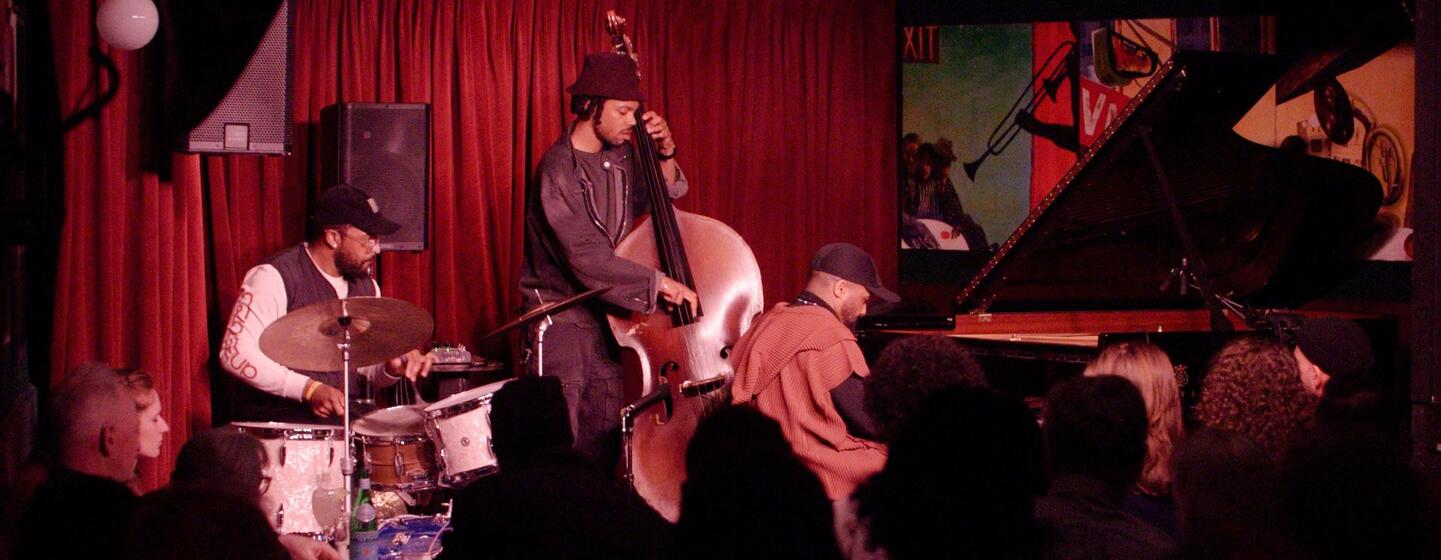 A jazz trio performing on stage at the Village Vanguard, featuring a bassist, a drummer, and a pianist, with an audience watching in the foreground.
