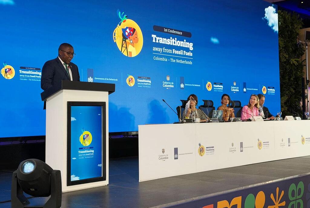 Selwin Hart speaking at a podium during the 1st Conference Transitioning away from Fossil Fuels in Santa Marta, Colombia. A panel of four people is seated at a long table behind him, with a large blue screen displaying the conference logo and title.