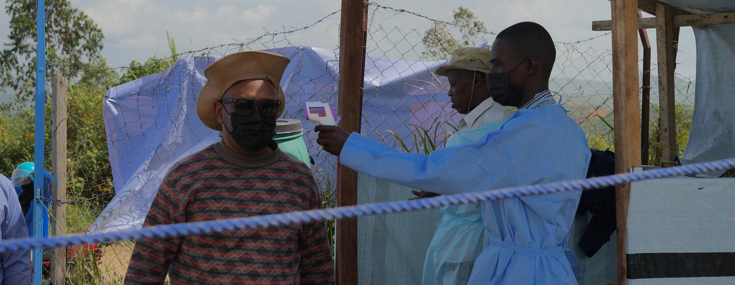 Un travailleur de santé en blouse bleue et masque utilise un thermomètre pour dépister un homme à l'entrée d'un établissement médical temporaire dans la province d'Ituri, en RDC.