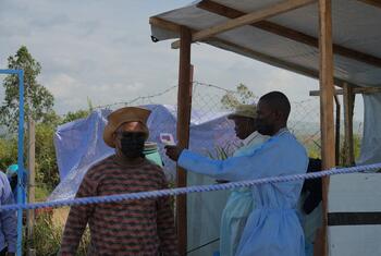 A health worker in a blue gown and mask uses a thermometer to screen a man at the entrance of a temporary medical facility in Ituri Province, DRC.