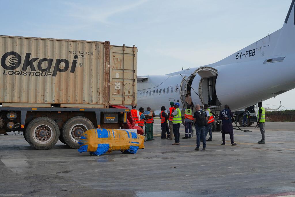 Wafanyakazi wenye mavazi ya usalama wanapakia kontena kubwa kutoka kwenye lori hadi kwenye ndege kwenye uwanja wa ndege. Kontena hiyo ina logo ya Okapi Logistique.